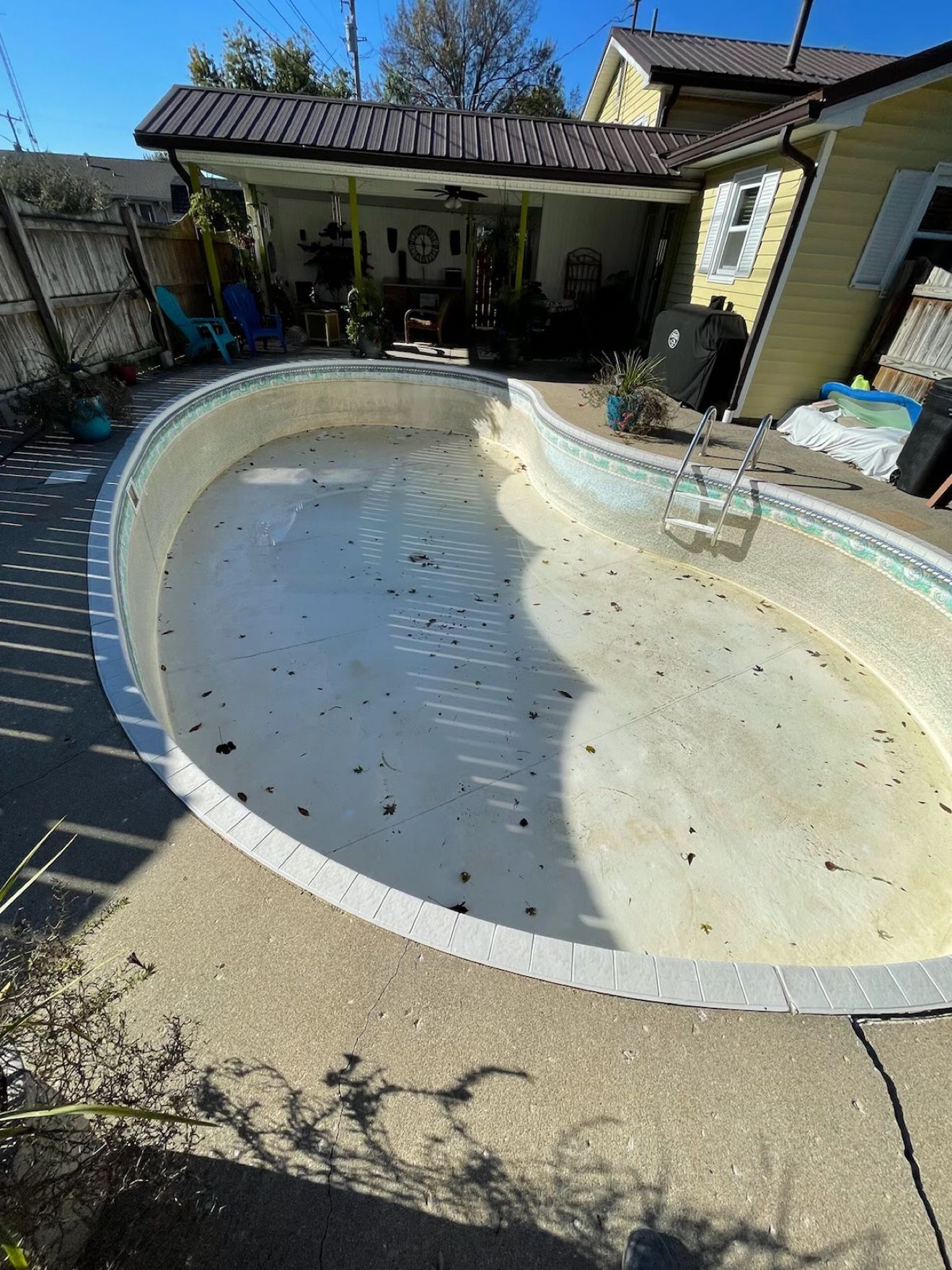 Kidney-shaped pool sitting empty before a liner replacement, faded liner with leaves in it