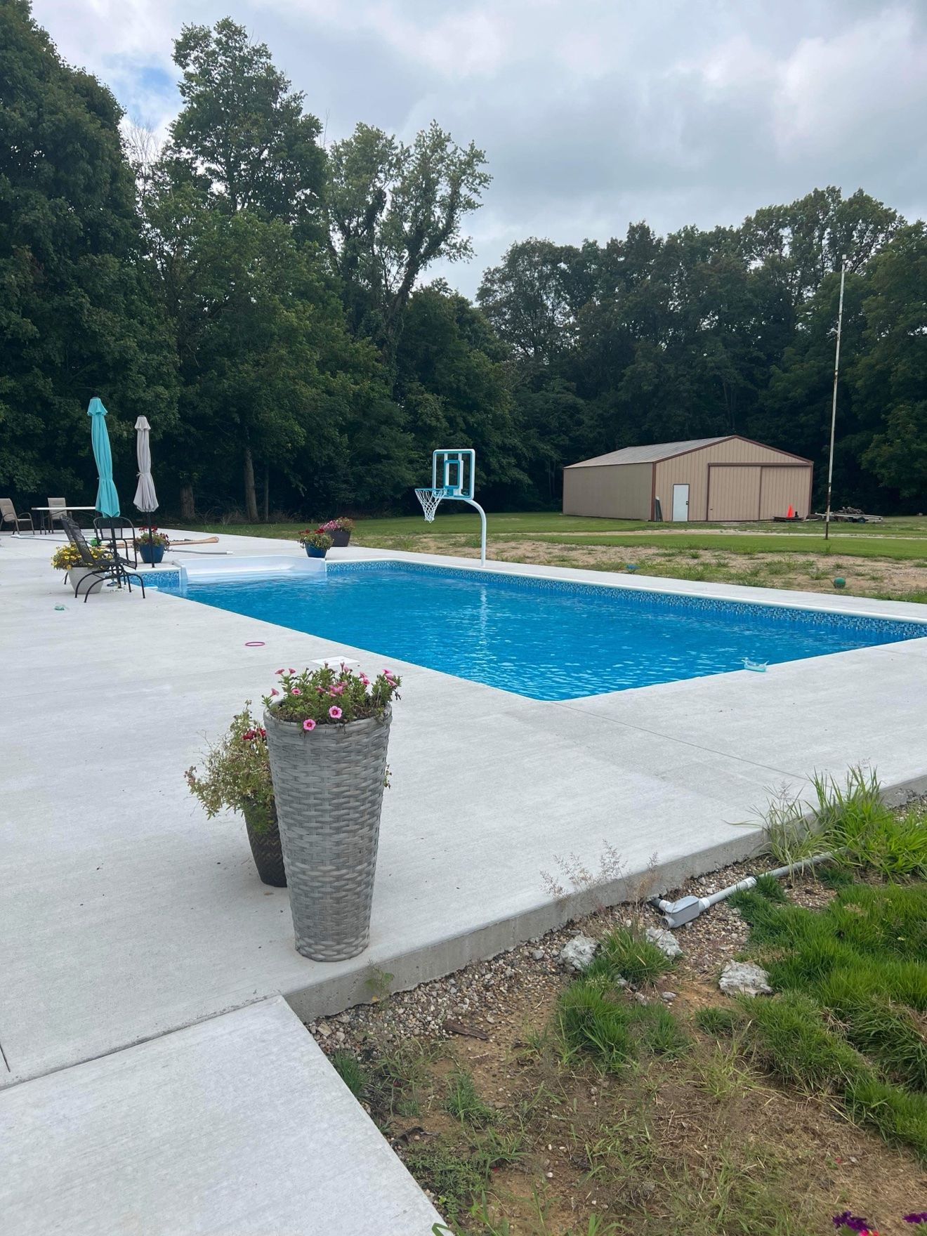 New inground pool build with bright blue water, a wide concrete deck, pool basketball hoop and flower planters